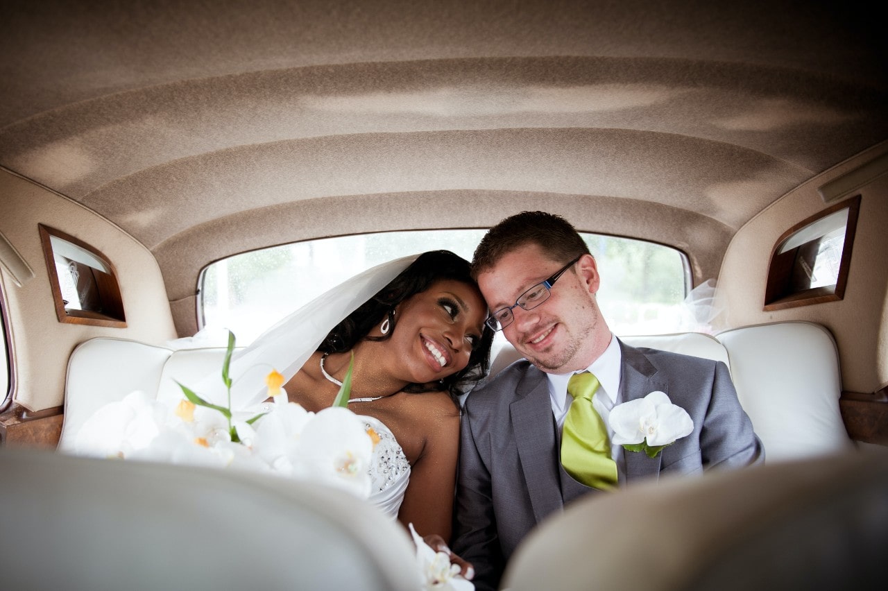 Bride and groom in the backseat of a car, resting their heads on one another’s and smiling