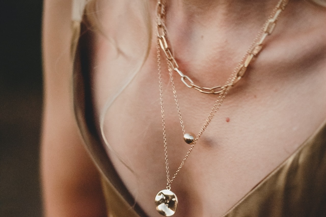 A woman wearing three yellow gold necklaces: a large gemstone pendant that’s longest, a shorter and smaller metal pendant, and then the shortest necklaces is a gold paperclip chain necklace
