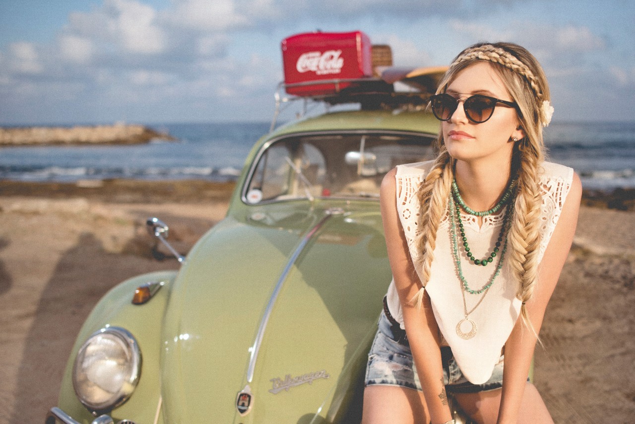 A woman at the beach sitting on the hood of her car wearing multiple beaded necklaces at different lengths with an even longer gold circle pendant necklace