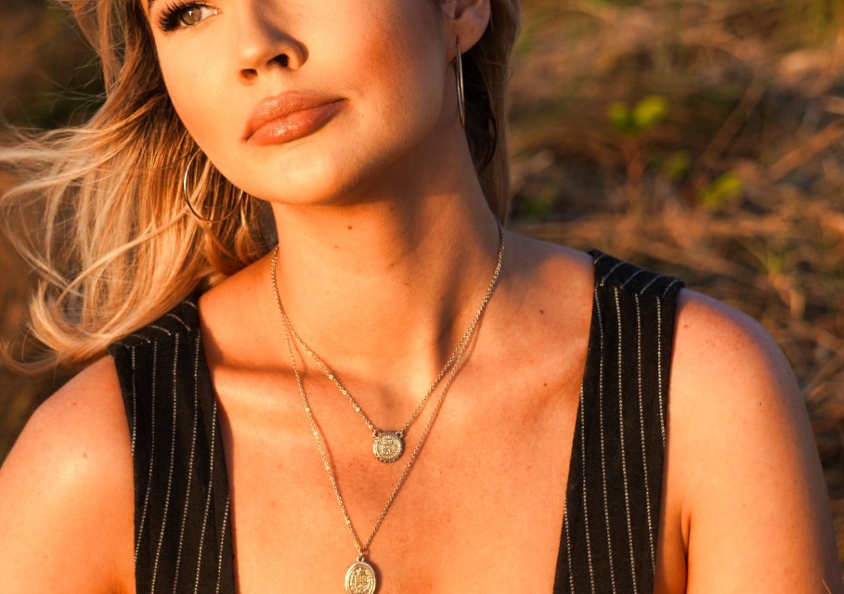 A woman watching the sunset while wearing two gold medallion necklaces, one on a shorter chain and smaller pendant than the other