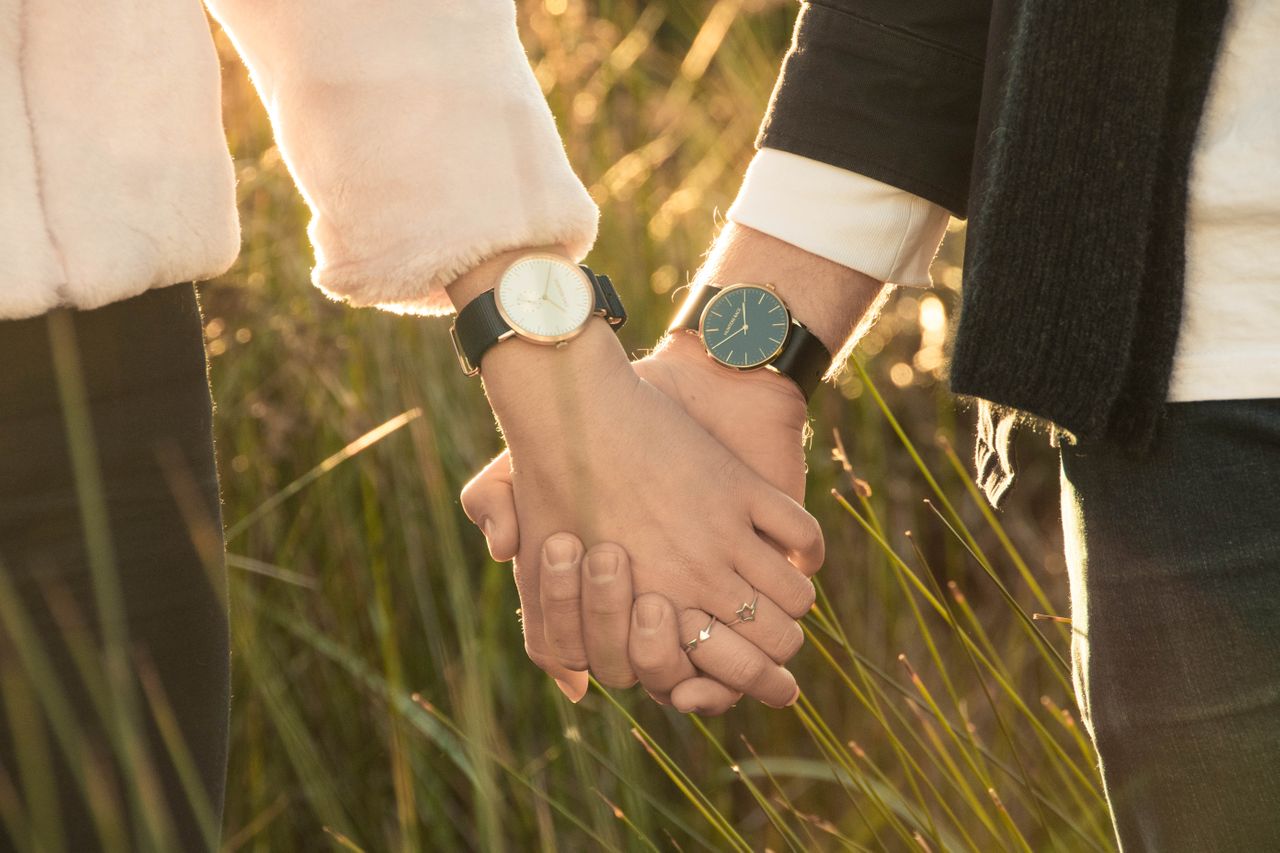 A timepiece-loving couple holds hands with matching watches while walking through an overgrown field at sunset