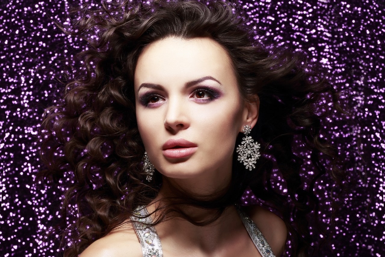 A woman wearing elaborate diamond earrings stands against a purple glitter background.