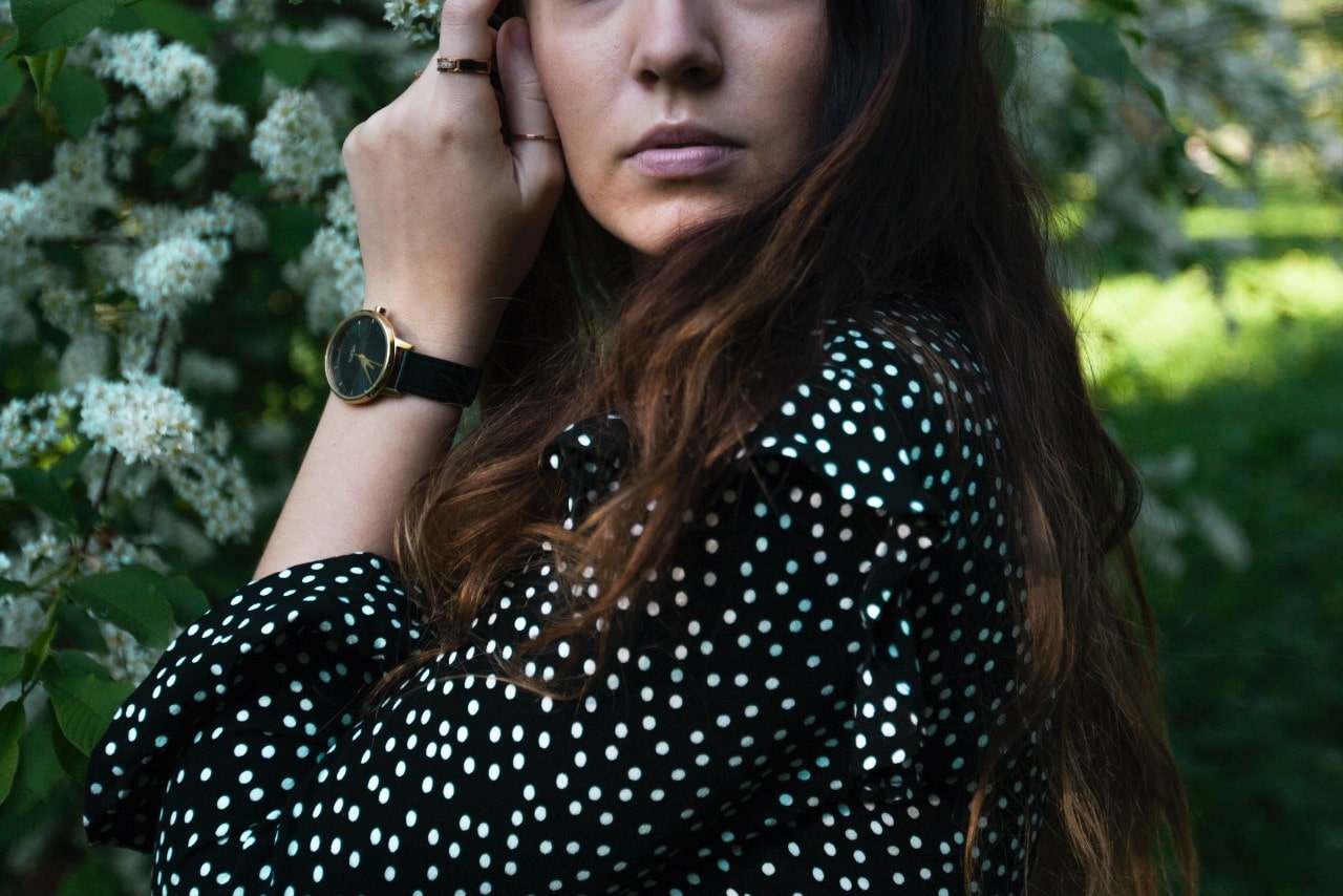 A woman wearing a polka-dotted shirt and luxury watch in front of delicate white flowers.