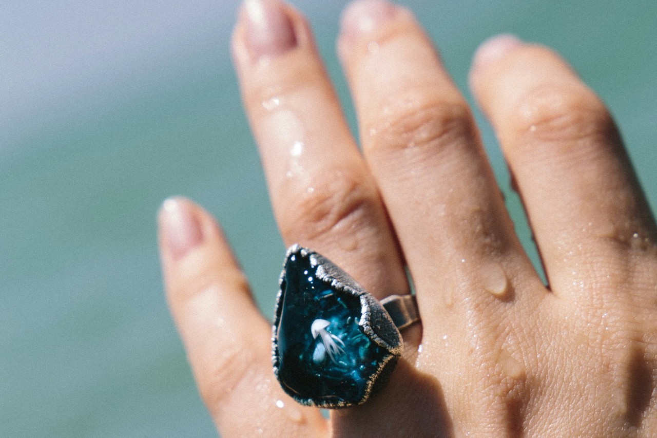 A person’s hand extended by the water, wearing a large blue gemstone ring