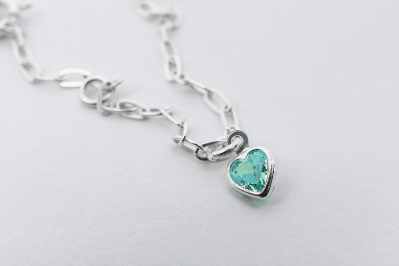 A white gold chain necklace with a heart pendant and a light blue gemstone.