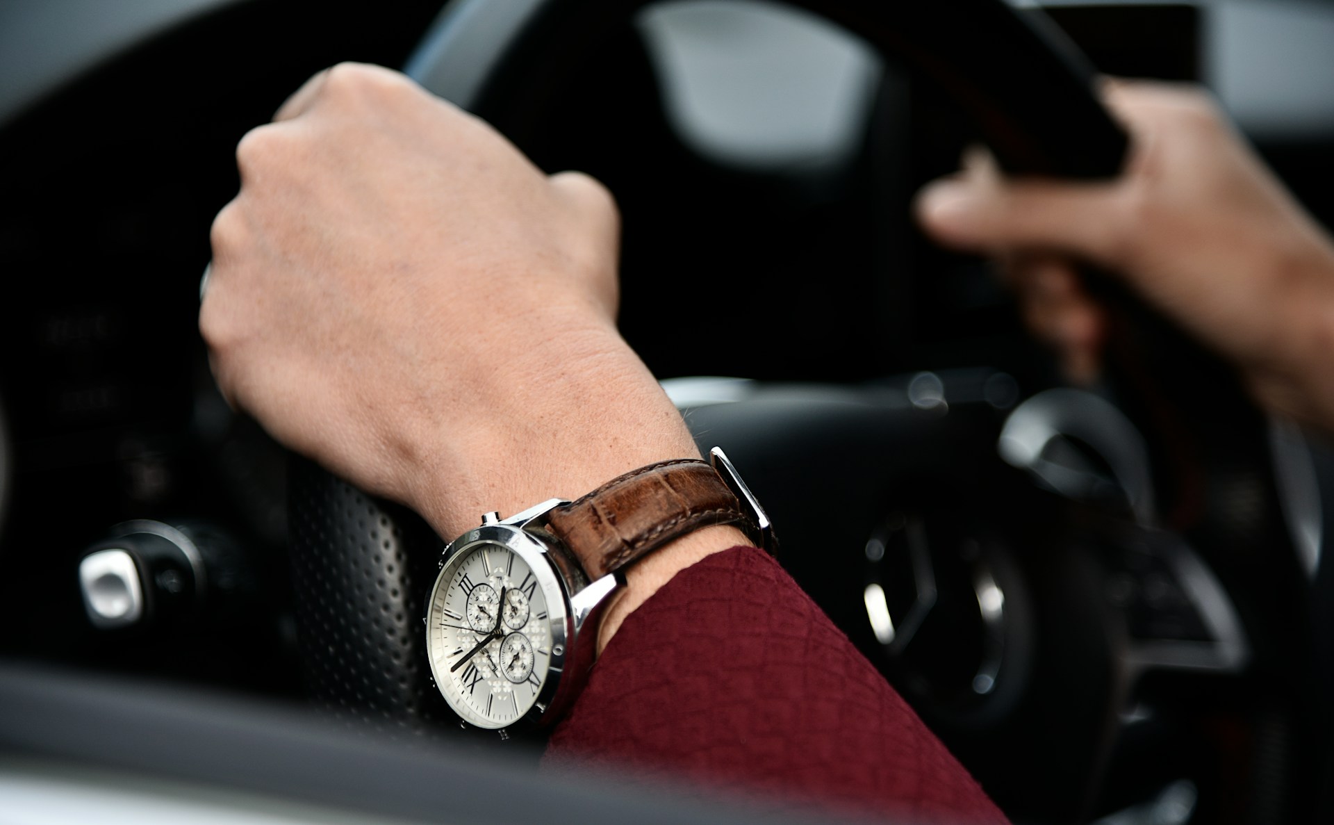 a pair of hands holding a steering wheel and adorned with a silver watch with leather strap