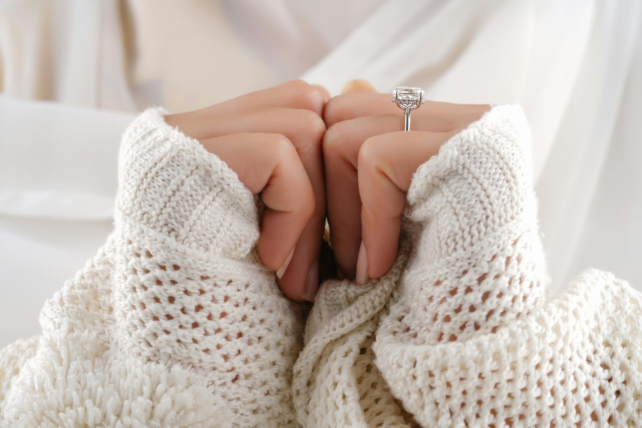 A close-up of a woman in a comfortable sweater, an elegant engagement ring on her finger.