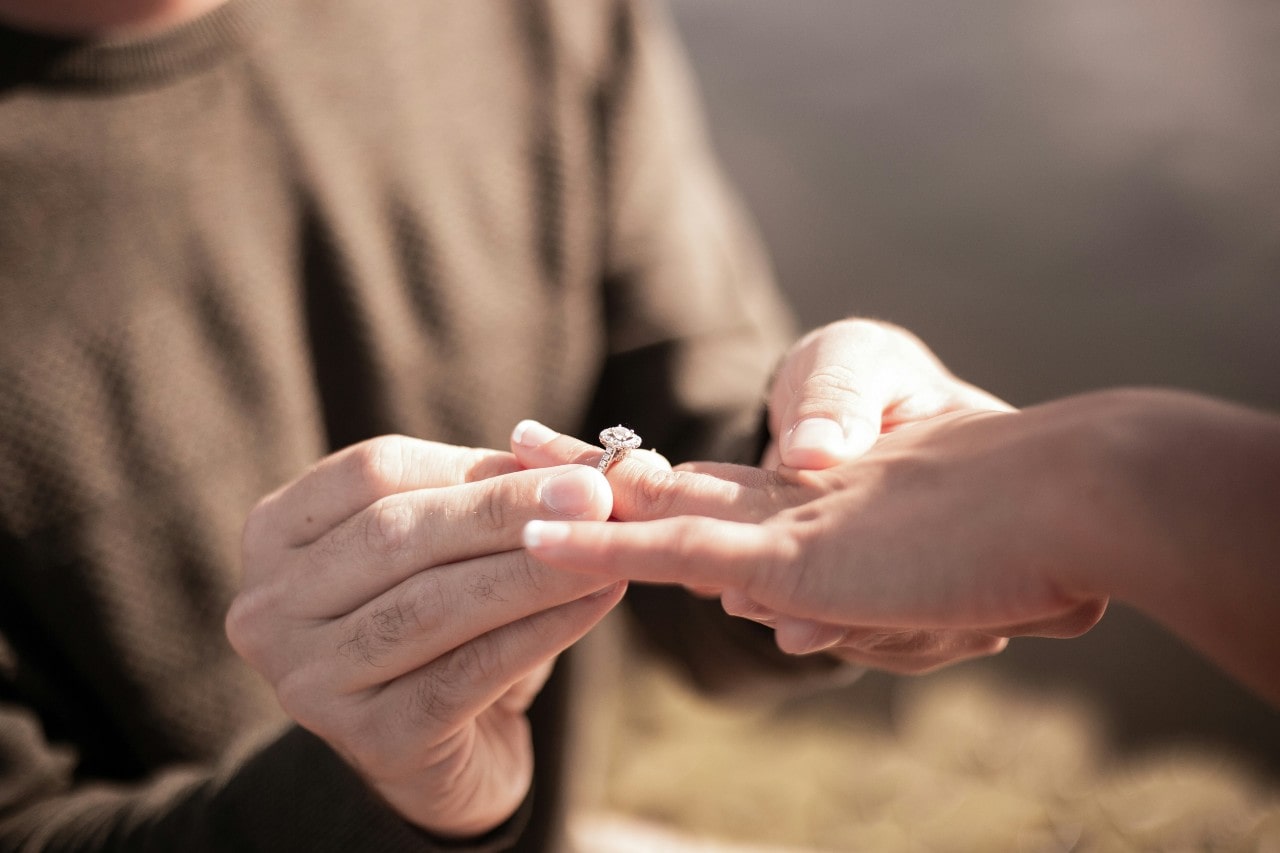 A close-up of a man putting an elegant halo engagement ring on his partner’s hand.