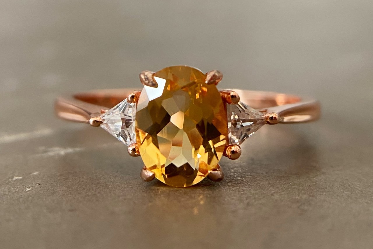 A stunning rose gold ring features an oval-cut yellow gemstone flanked by two sparkling white diamonds, set against a neutral background