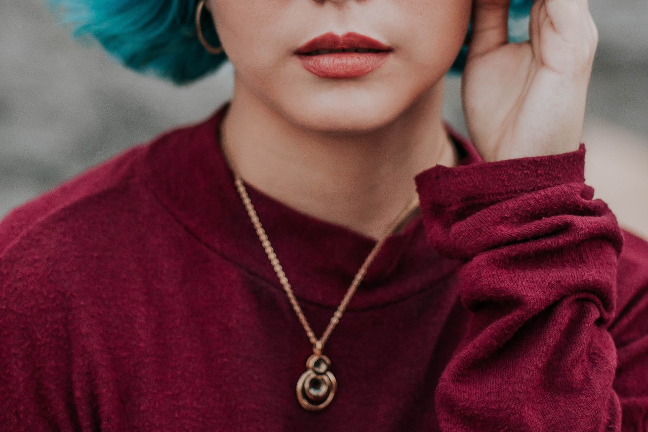  rose gold pendant necklace with a circular design rests elegantly on the collarbone of a woman wearing a deep red sweater and blue-tinted hair