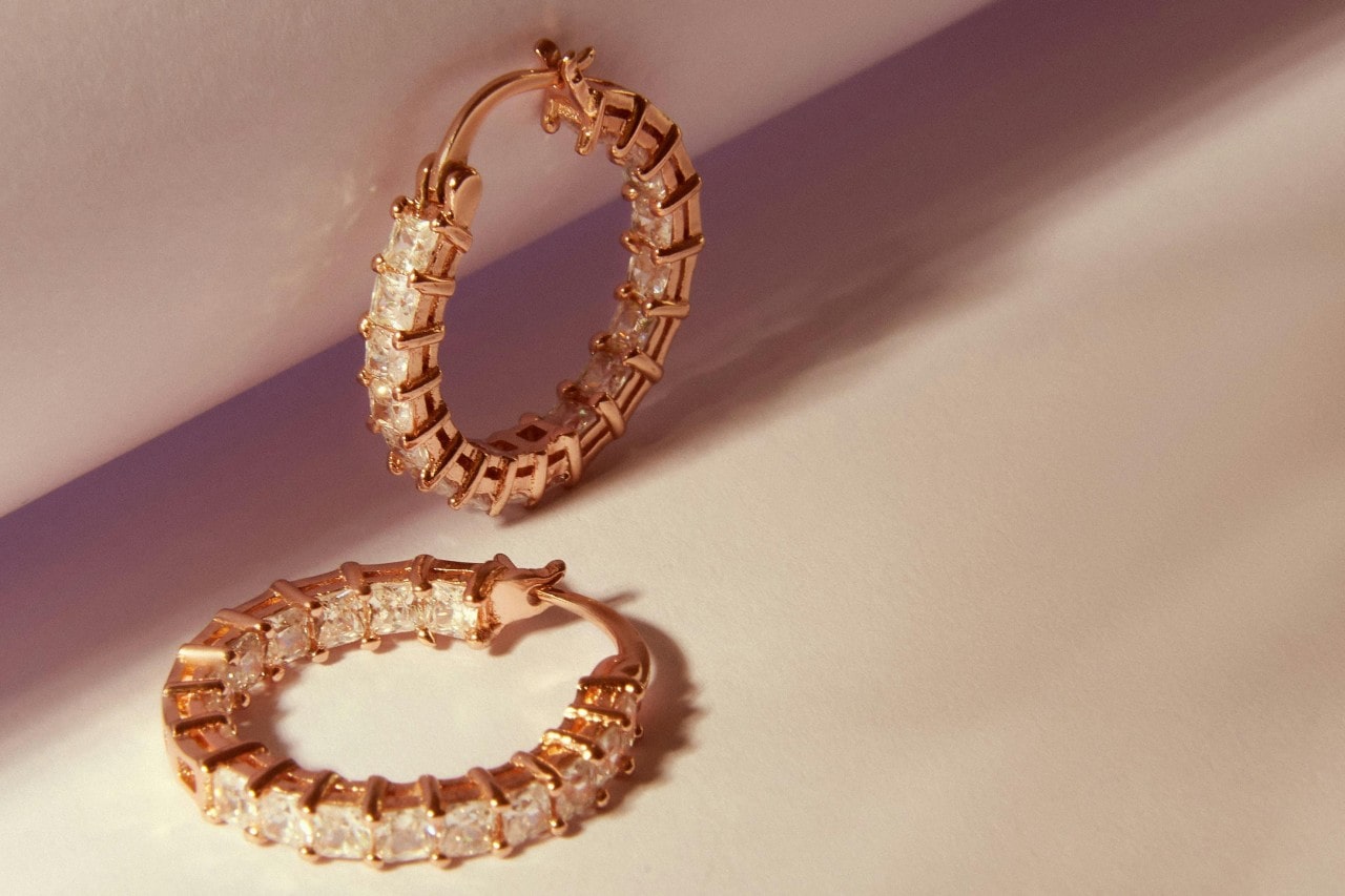 A pair of rose gold hoop earrings featuring a continuous row of radiant-cut diamonds on a pastel pink background.