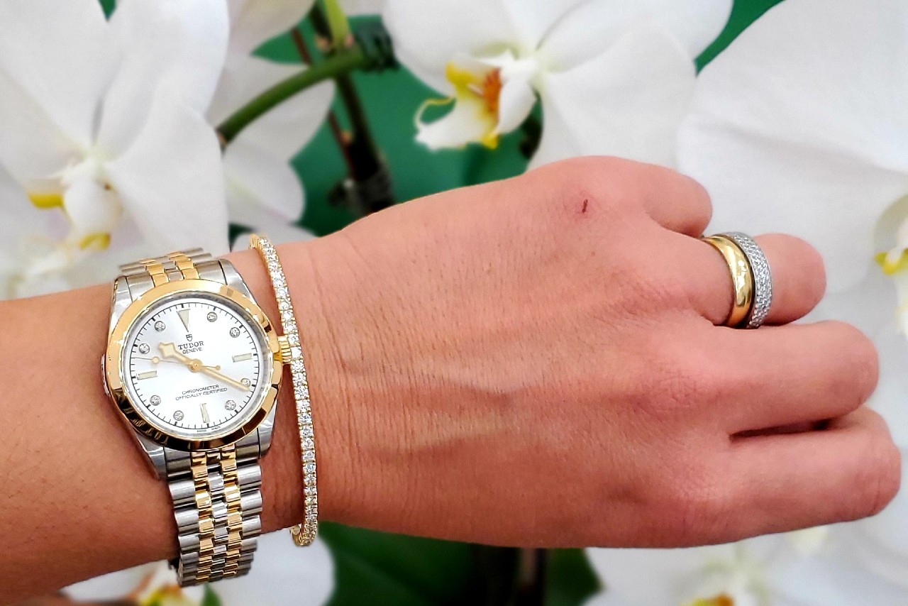a diamond bracelet next to a TUDOR mixed metal watch on a woman’s wrist along with a yellow gold and white gold diamond band stacked on top of each other on the woman’s hand